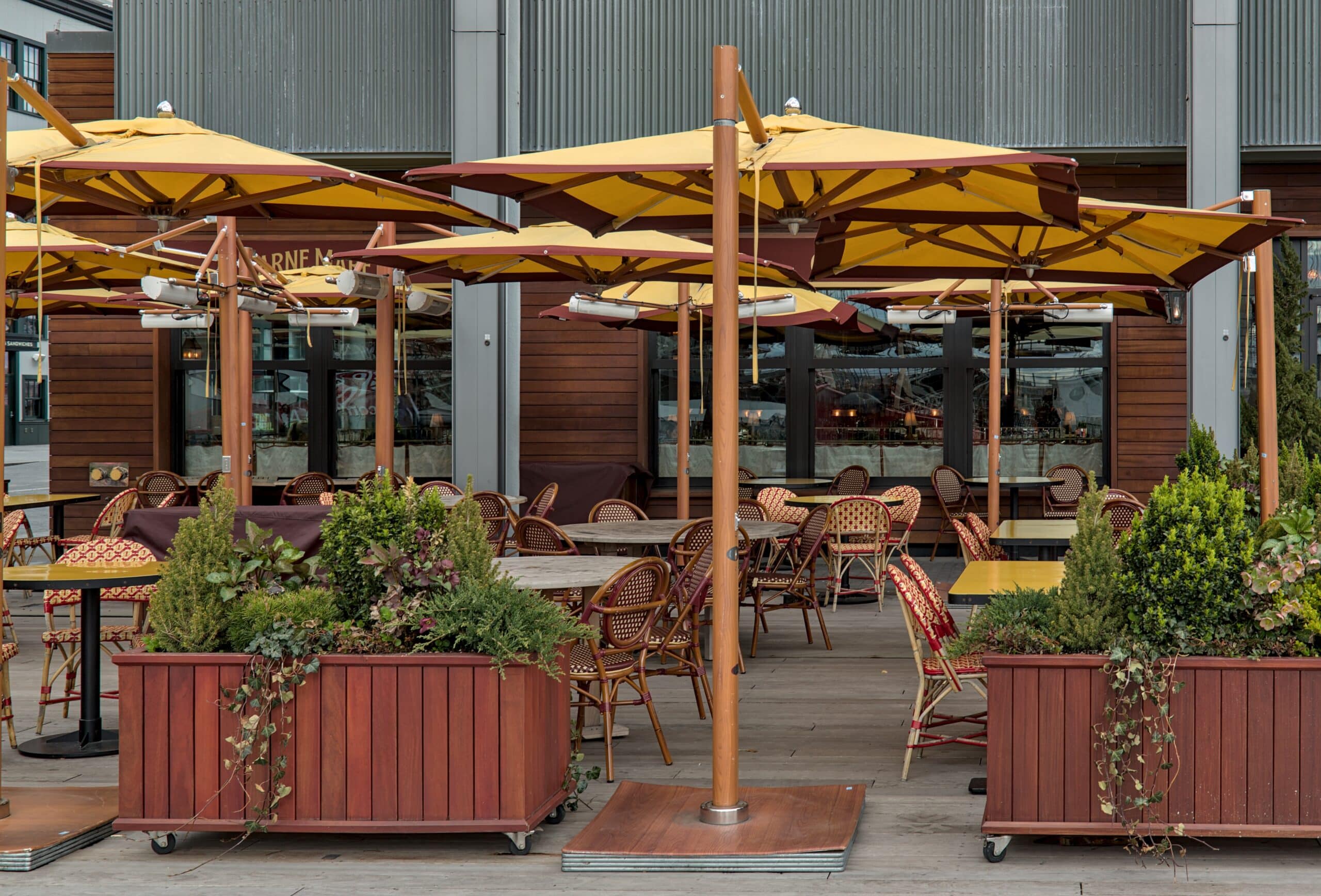 Cozy restaurant patio with elegant wooden tables and chairs, shaded by large umbrellas. The space is adorned with potted plants, adding a touch of greenery. Soft, warm lighting enhances the inviting atmosphere, perfect for outdoor dining