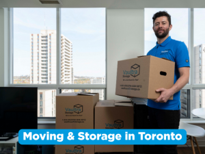 A Vaultra Storage employee in a blue polo shirt holds a branded cardboard moving box inside a bright, high-rise apartment or office in Toronto, with additional Vaultra-branded boxes stacked on the floor and a city skyline visible through floor-to-ceiling windows. A bold banner at the bottom reads "Moving & Storage in Toronto."